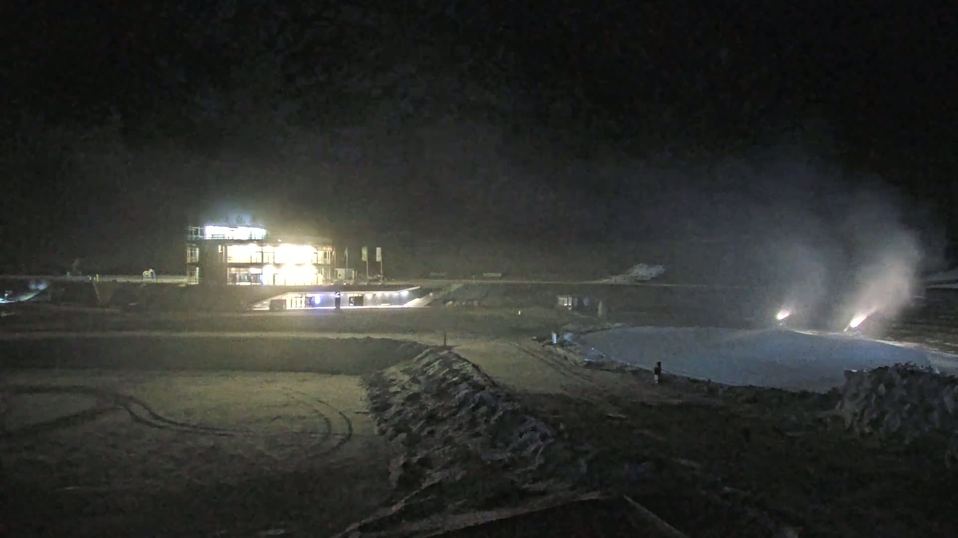 At night, snow cannons create plumes of snow on a snow-covered surface near a brightly lit building.