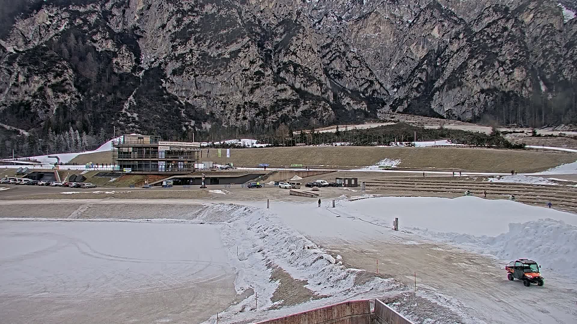 A partially snow-covered outdoor sports complex sits at the base of a large mountain range on a cloudy day.