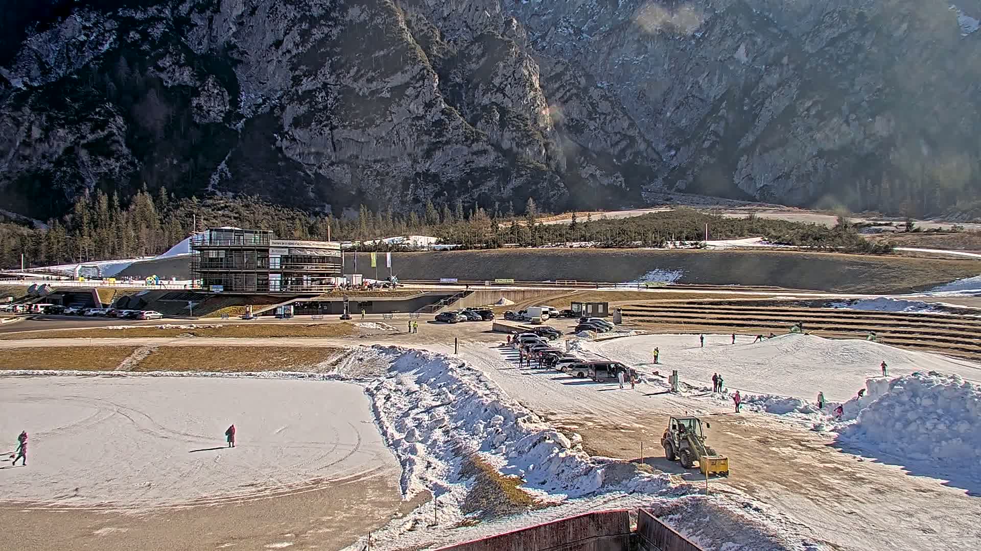 A sunny day at a winter sports facility nestled in a mountainous valley, featuring a modern building, snow-covered areas, parked cars, and people enjoying winter activities.