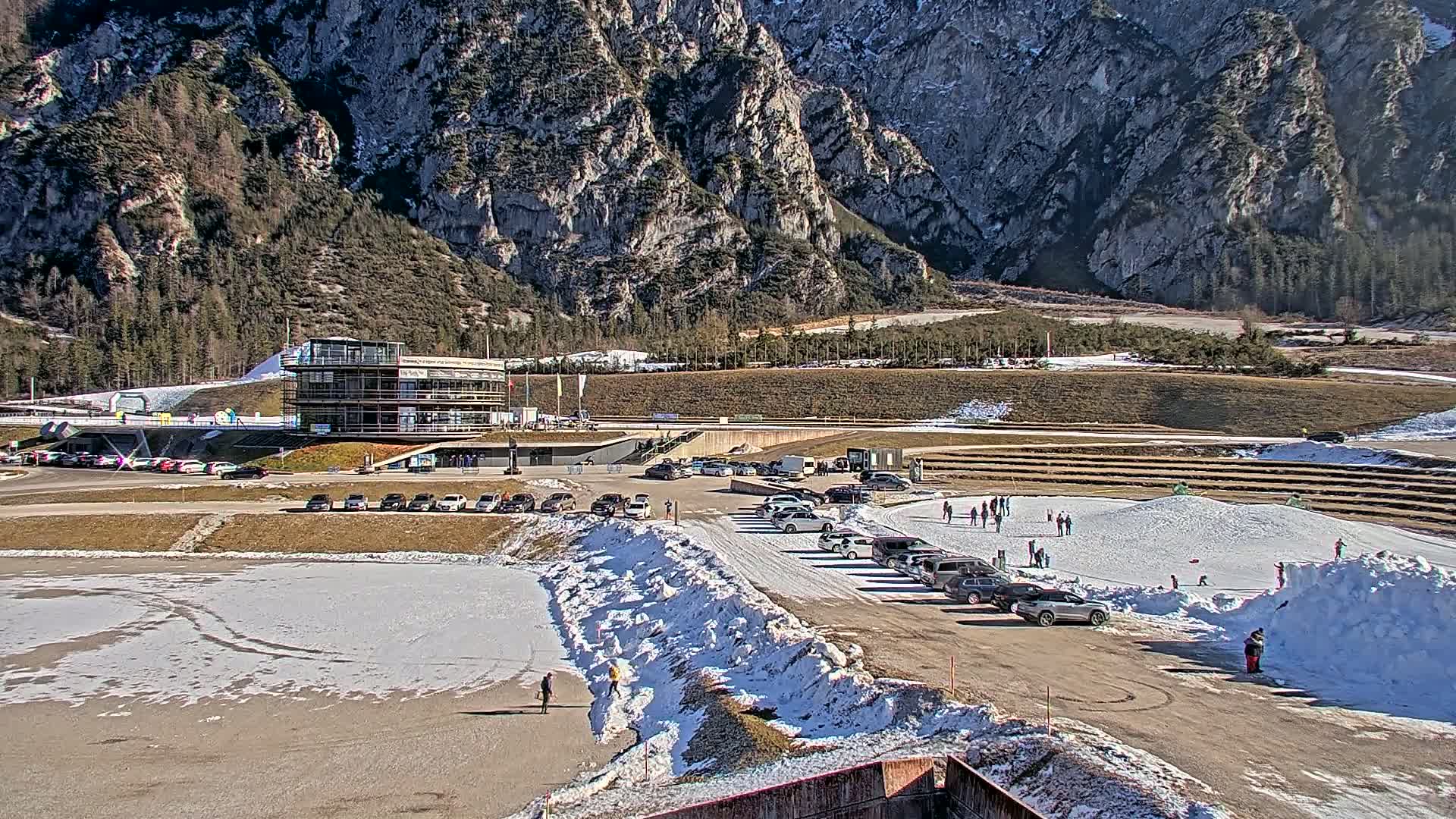 A snowy mountainside sports complex with a multi-story building, parking lots filled with cars, and people enjoying snowy areas, under a sunny sky.