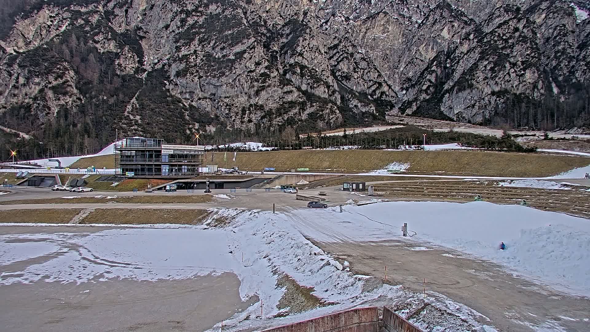 A multi-story building sits in a snowy parking lot next to a partially snow-covered dirt track, with a large mountain in the background.