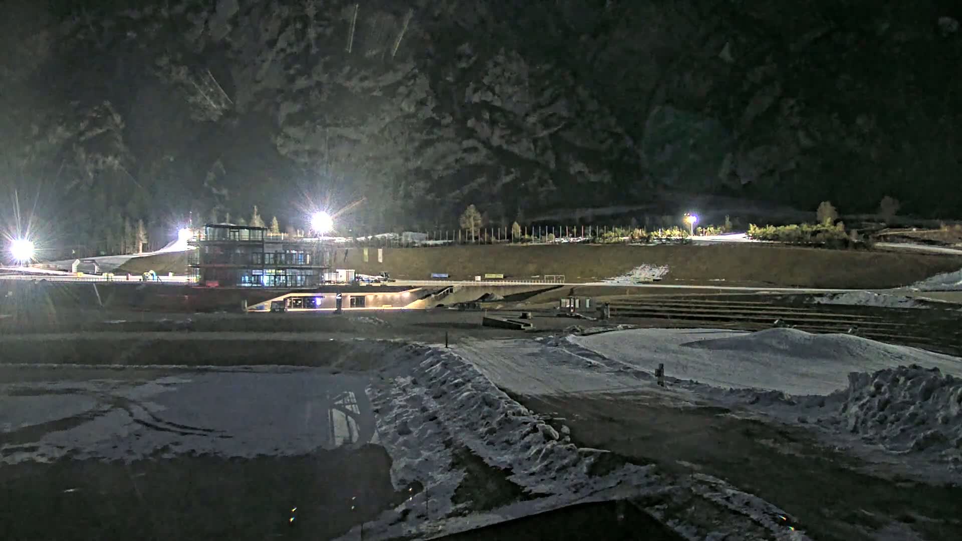 A nighttime view of a snow-covered outdoor sports complex with a modern building illuminated by bright lights against a dark mountain backdrop.
