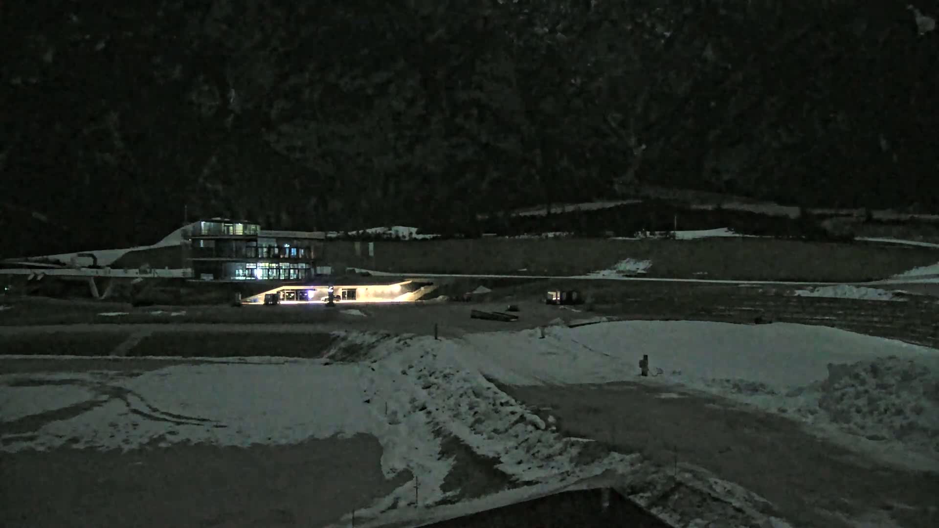A modern building with lights on is situated in a snowy, nighttime landscape against a dark mountain backdrop.
