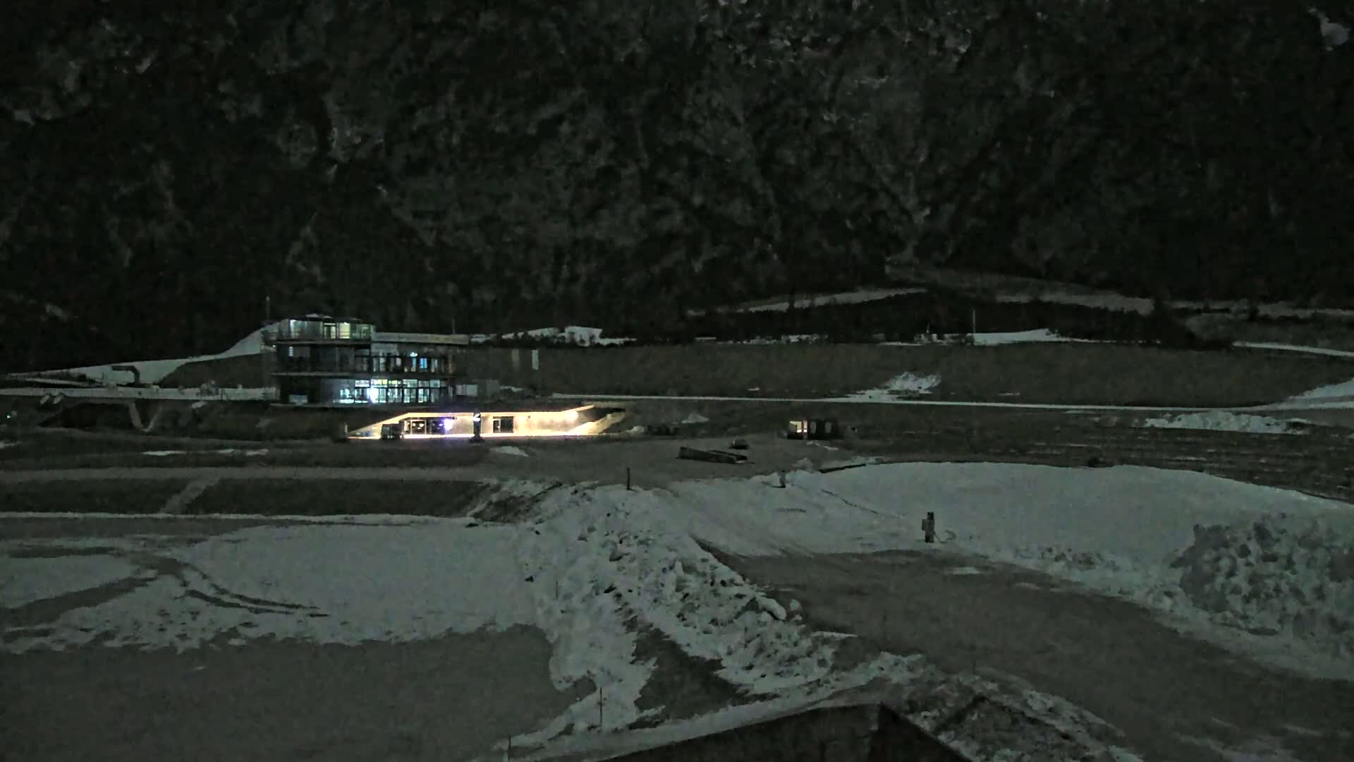 A brightly lit building sits on a snow-covered landscape at night, with a dark mountain range in the background.