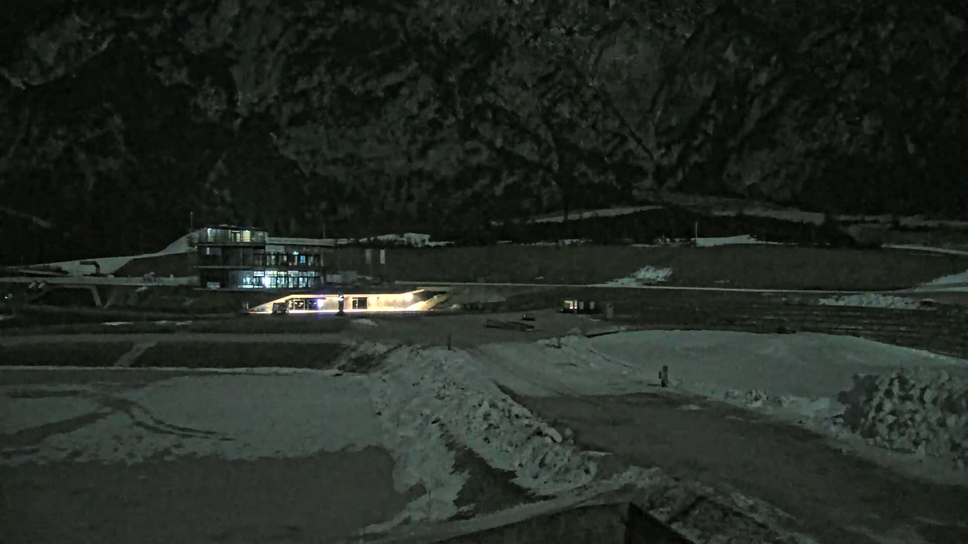A snow-covered, nighttime landscape features a lit building near a dark mountain range.