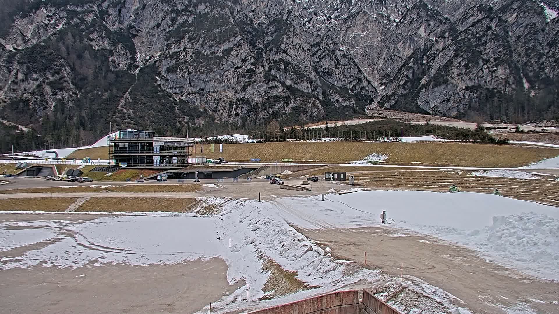 A multi-story building sits in a snowy, mountainous area with patches of dirt and some vehicles parked nearby.