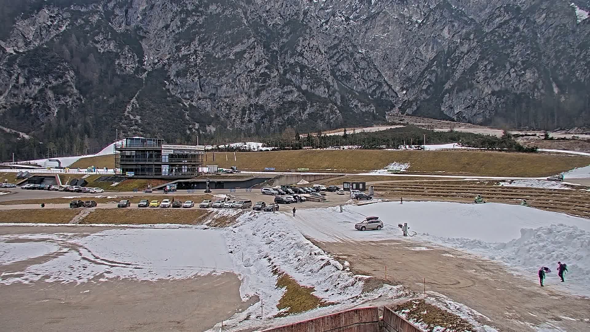 A parking lot with cars is situated below a modern building at the base of a large, rocky mountain, with patches of snow on the ground.
