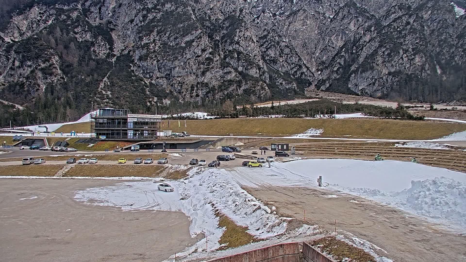 A parking lot with several cars sits in front of a multi-story building near a mountainside with patchy snow on the ground, under a cloudy sky.