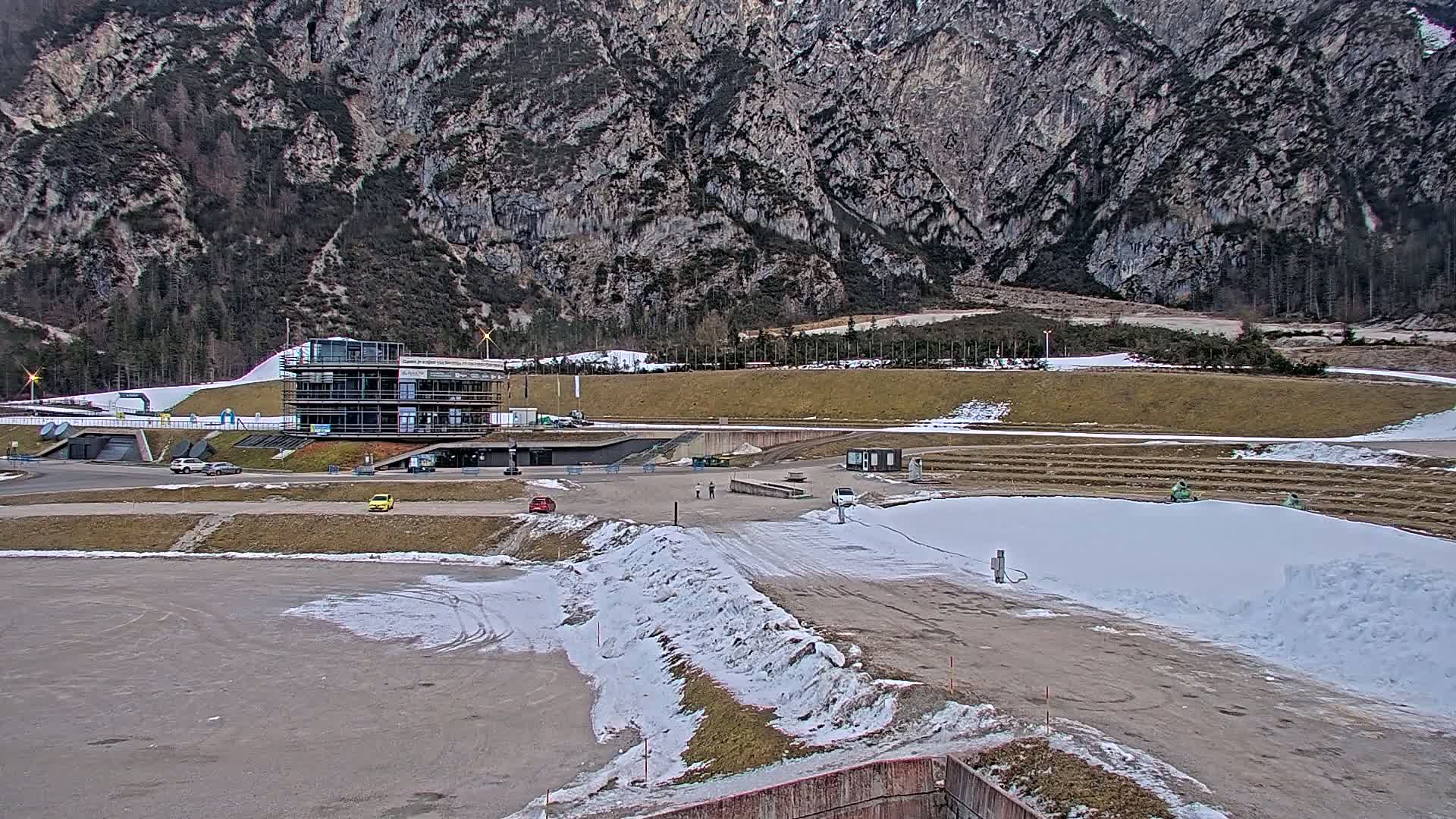 A modern building sits in a snowy, mountainous area with patches of bare ground and a few vehicles parked nearby.