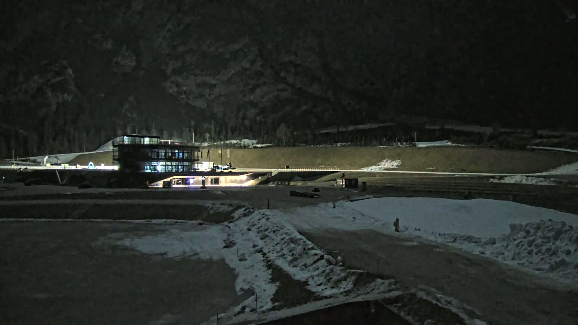 A snow-covered nighttime scene shows a lit-up building near a road, with mountains in the background.