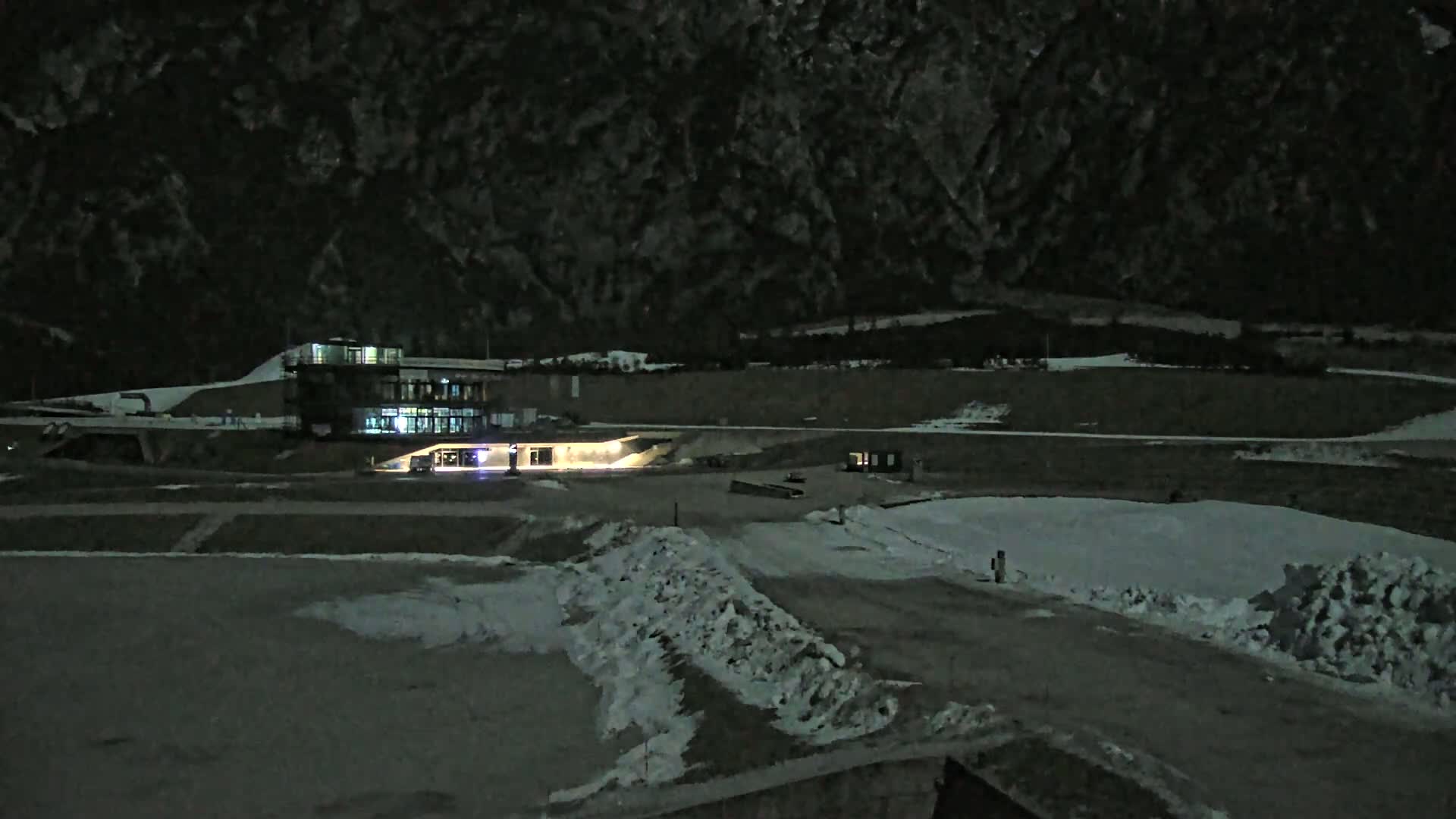 A nighttime, snow-covered landscape features a lit building near a mountain under a dark sky.
