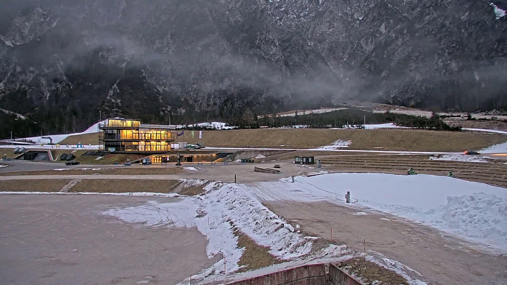 A multi-story illuminated building sits in a snow-dusted, mountainous area under a cloudy, overcast sky.