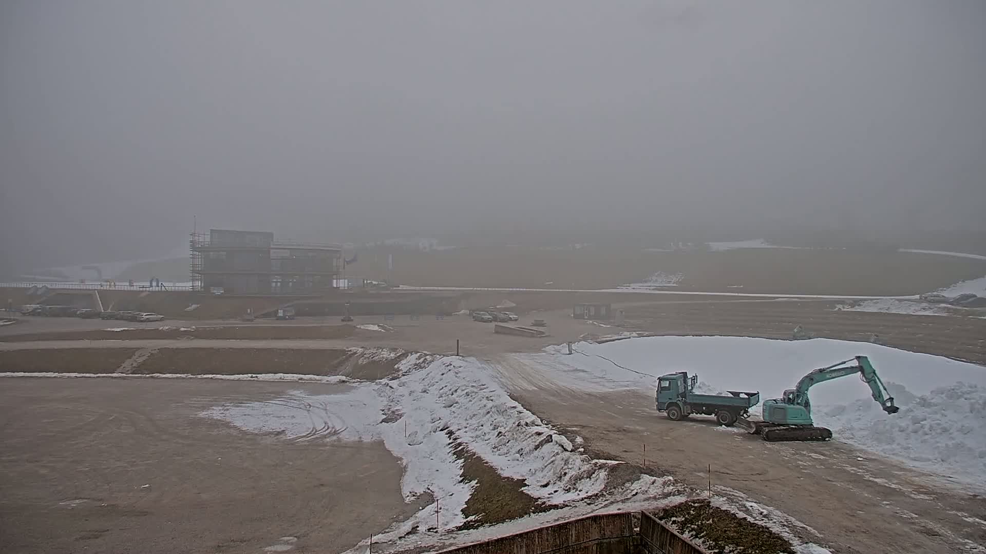 A foggy construction site with a partially built structure, several vehicles, and piles of snow.
