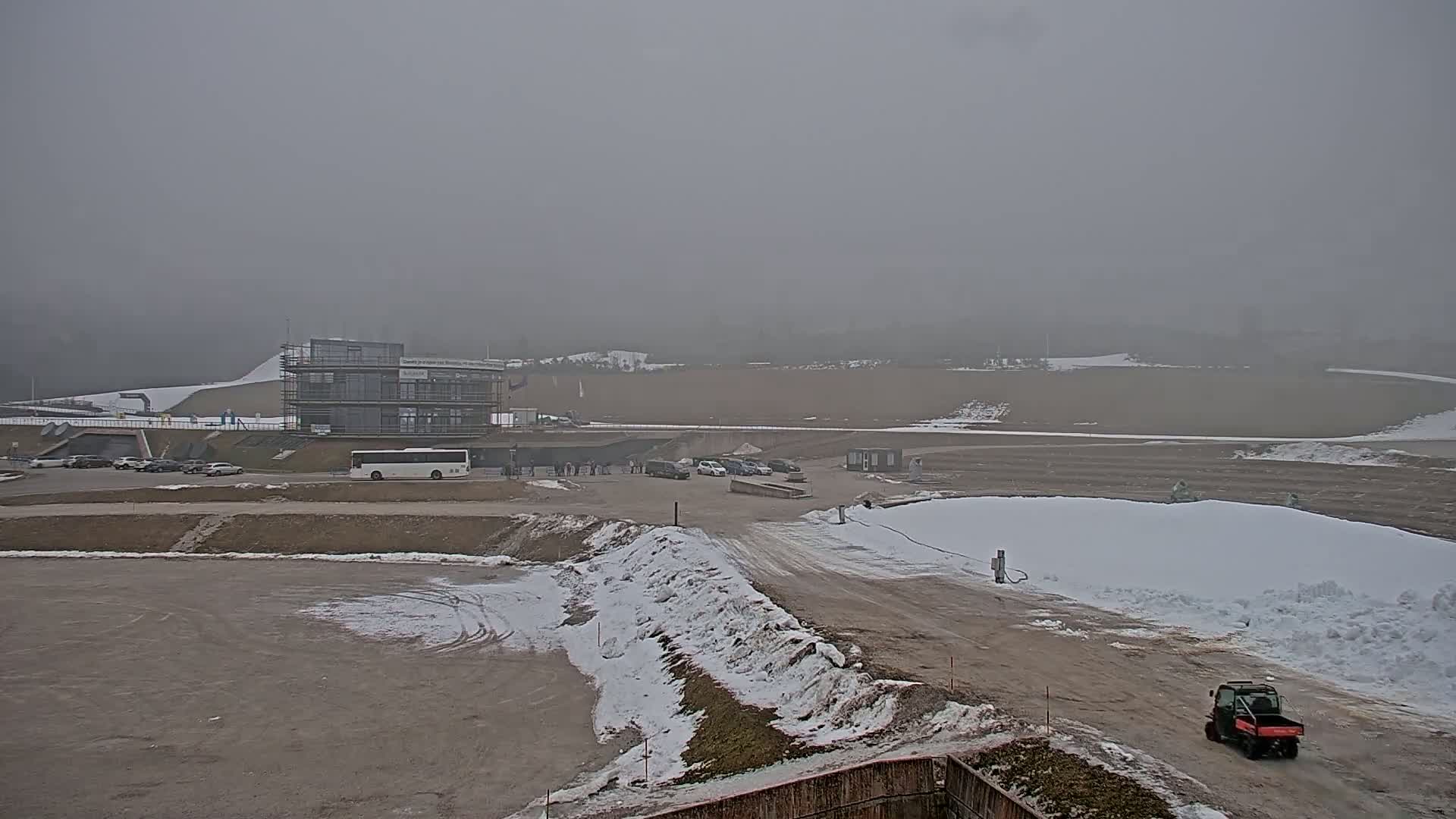 A foggy, snowy landscape shows a parking lot with several vehicles near a partially constructed building, with snow piles and dirt areas visible.