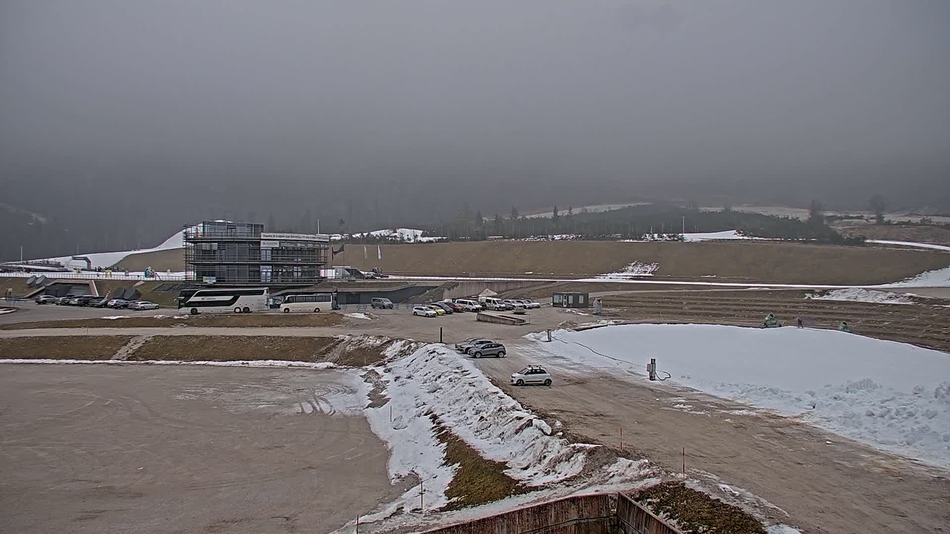 A parking lot with several cars and buses sits in front of a multi-story building, all of which are partially covered in snow under an overcast sky.