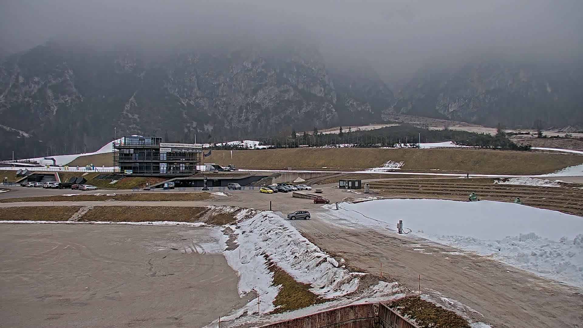 A large, mostly-empty parking lot and a modern building sit before a snow-dusted landscape and a mountain range shrouded in fog.