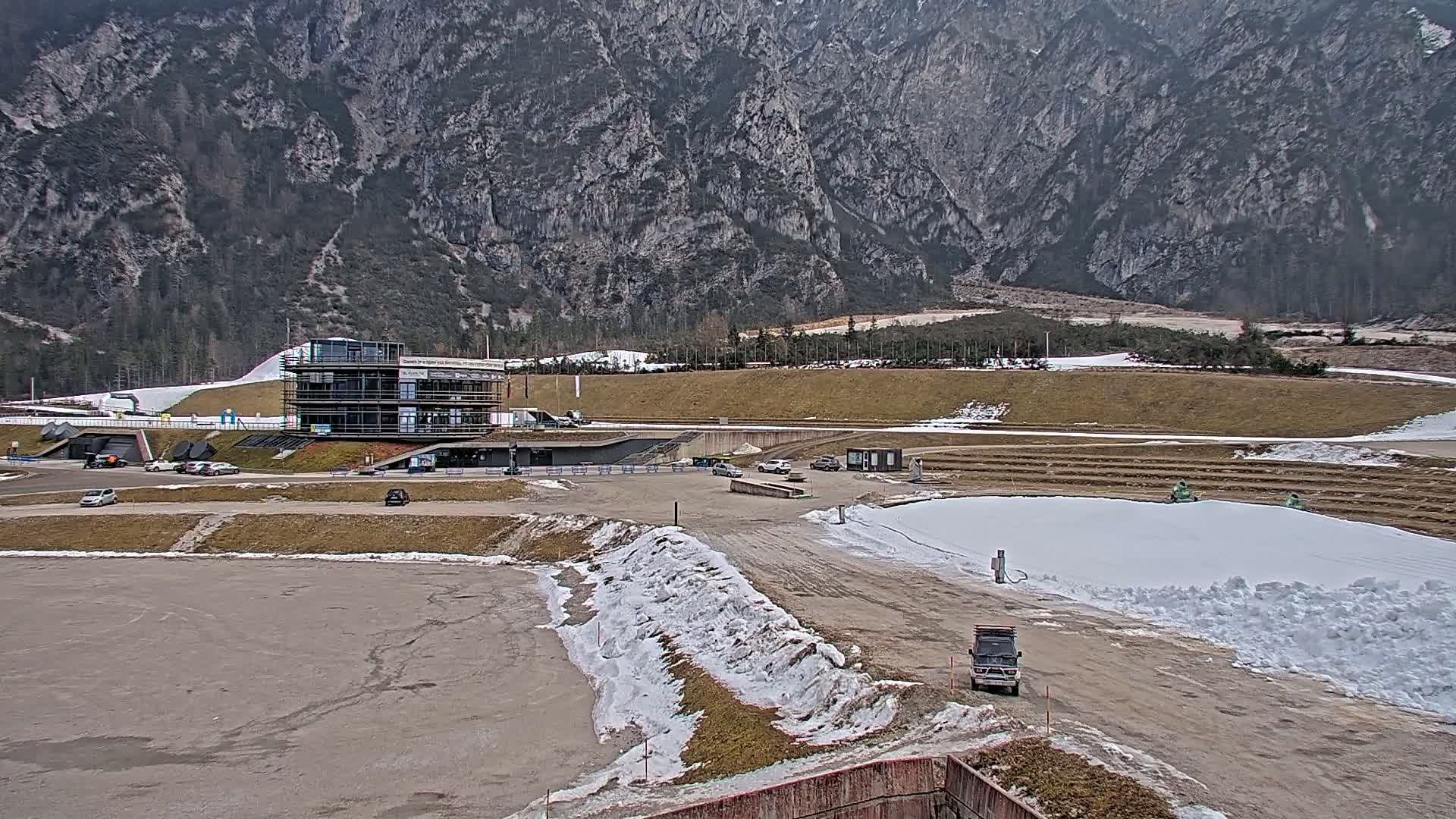 A modern building sits in a mountain valley with patches of snow on the ground, and a large, rocky mountain in the background under an overcast sky.