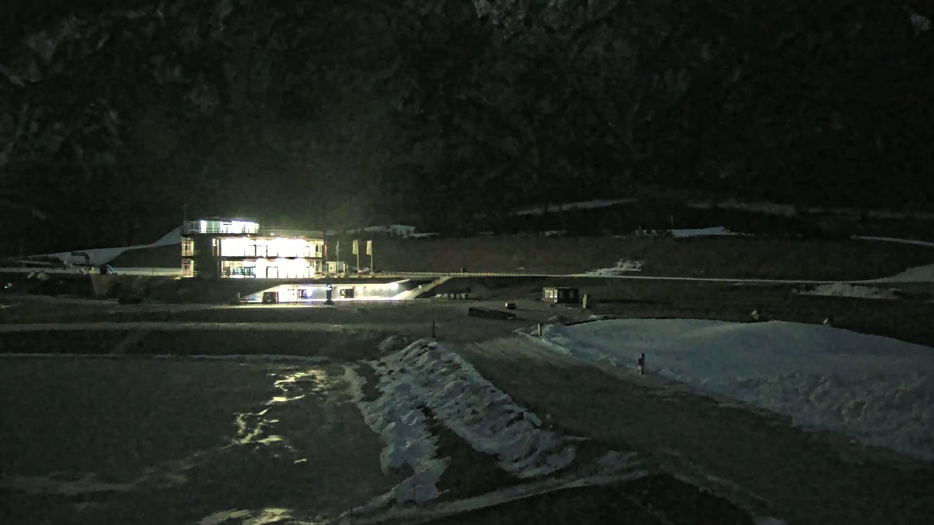 A brightly lit building stands in a snow-covered area at night, surrounded by dark hills under a clear sky.