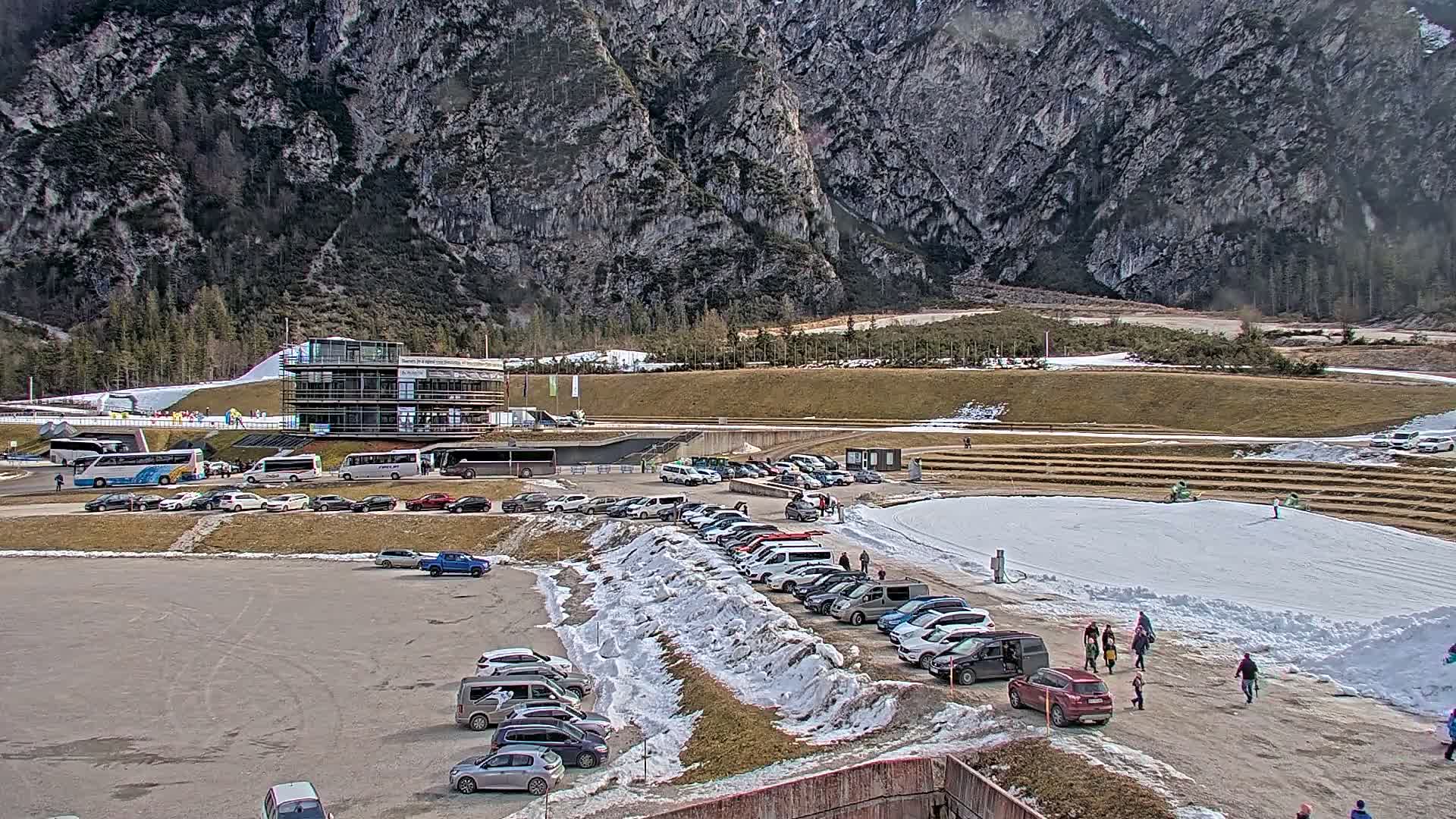 A large parking lot filled with cars sits below a multi-story building near a snowy area in front of a mountain range on a partly sunny day.