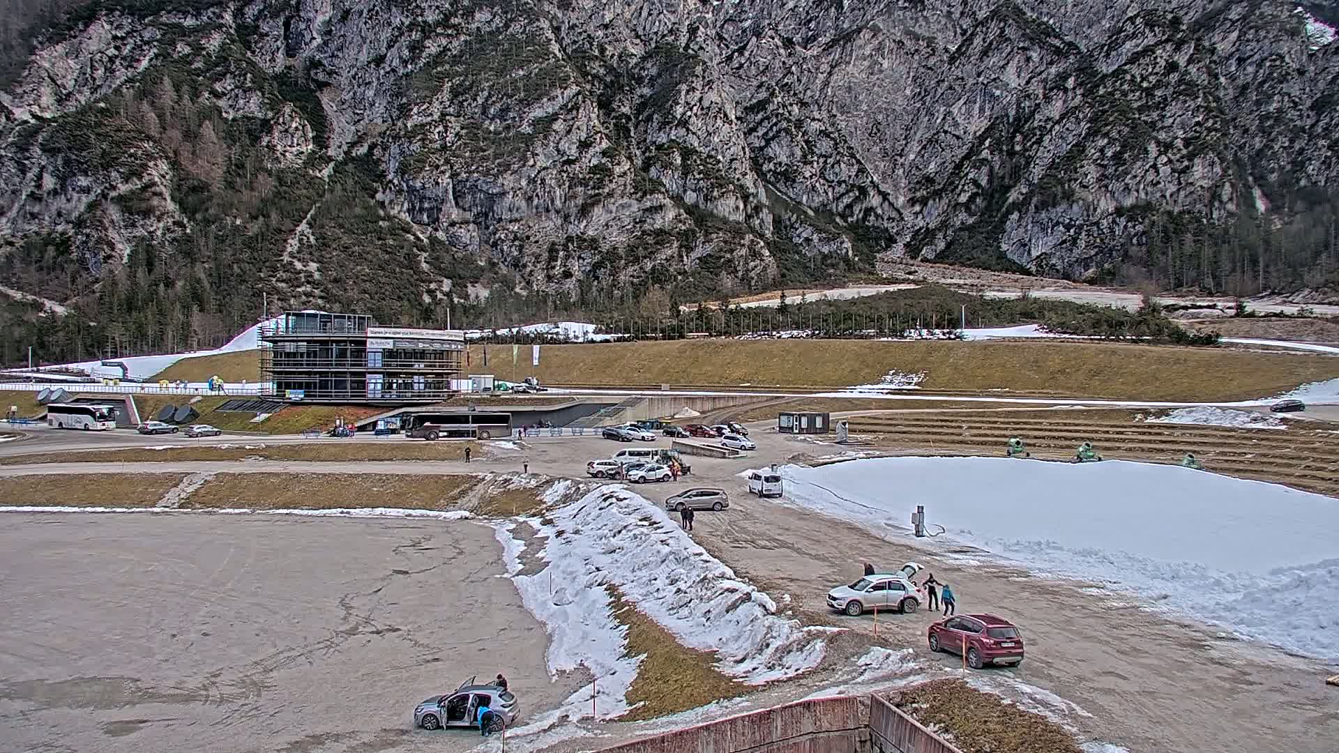 A parking lot with several cars and a multi-story building sits at the base of a large, rocky mountain under a mostly clear sky, with patches of snow and brown ground visible.
