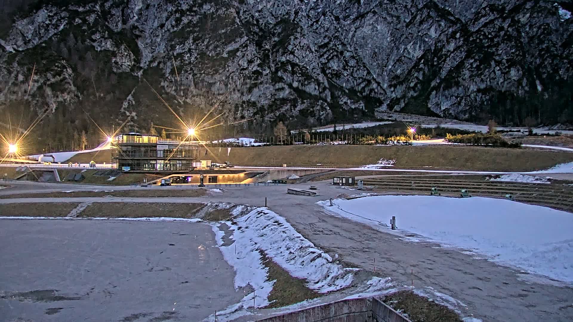 A nighttime view of a snow-covered outdoor sports complex nestled against a large, dark mountain, illuminated by artificial lights.