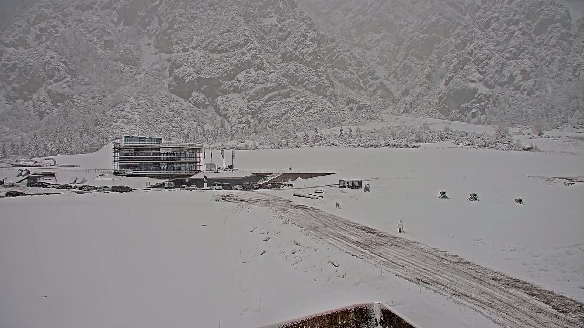 A snow-covered landscape with a multi-story building and a road leading to it, set against a backdrop of snow-laden mountains under a gray, overcast sky.