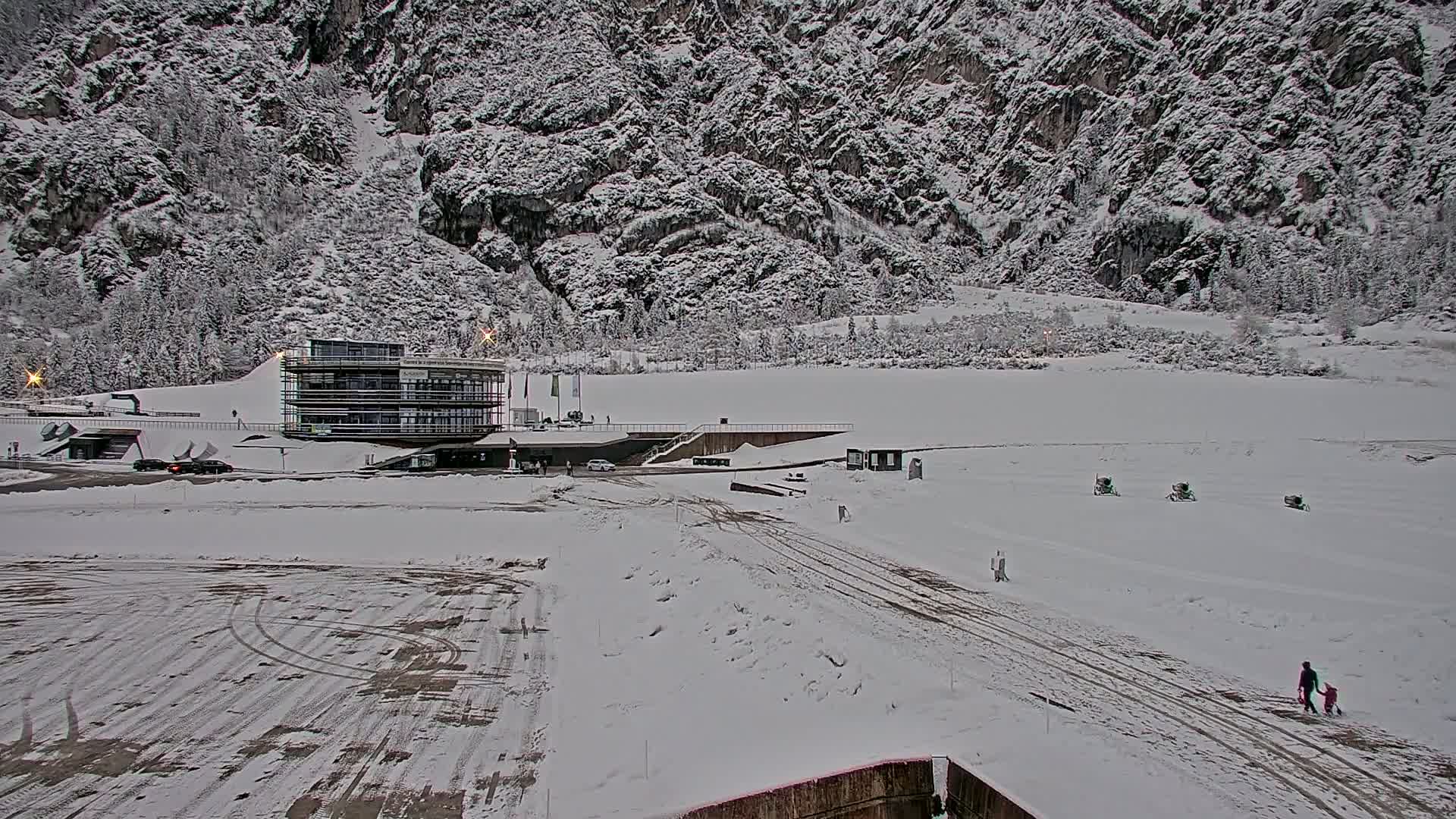 A snow-covered landscape features a modern building near a large, snow-covered flat area, with a mountainous backdrop under a cloudy sky.