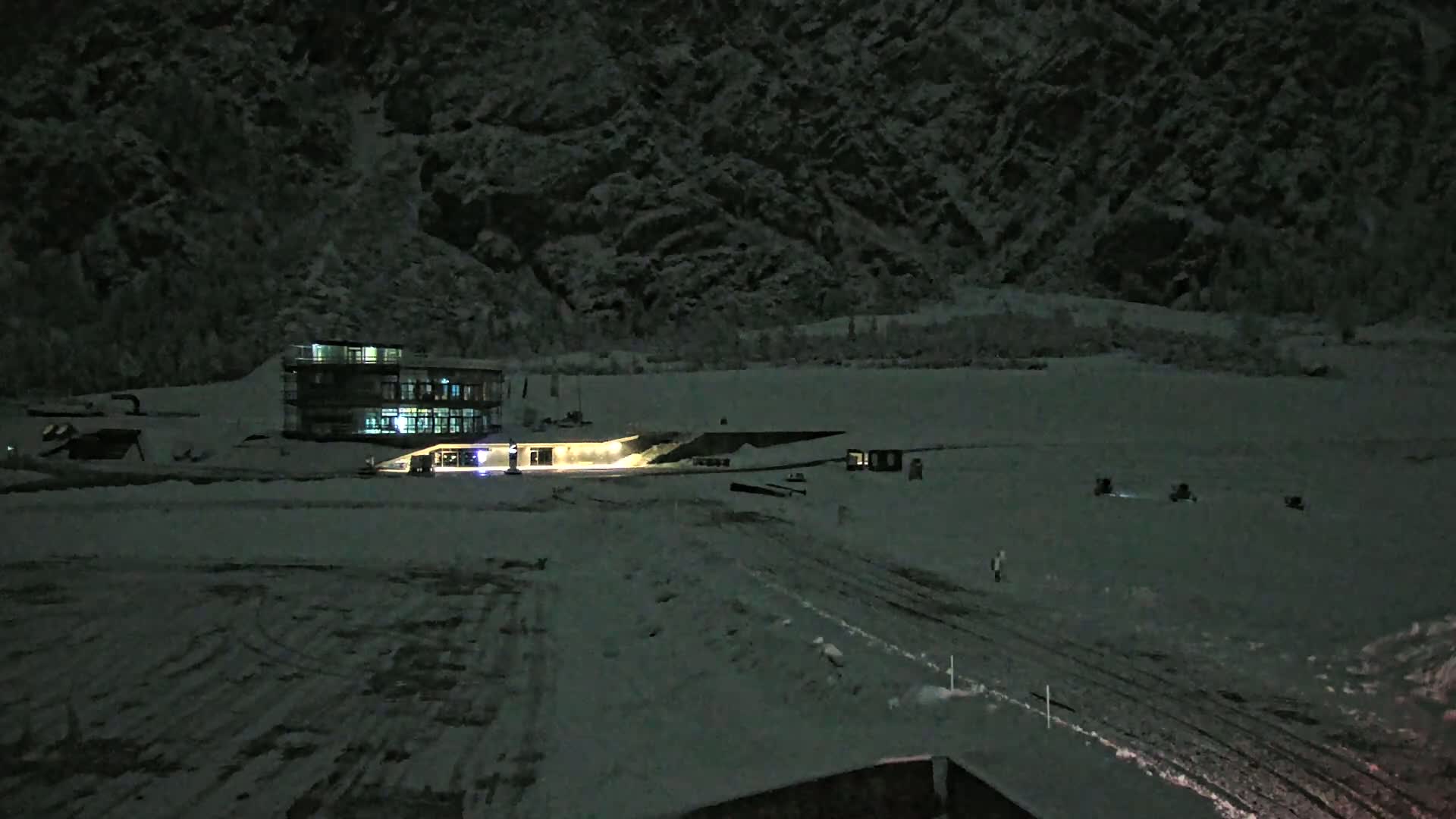 A snow-covered landscape at night, featuring a lit-up building near a dark mountainside.