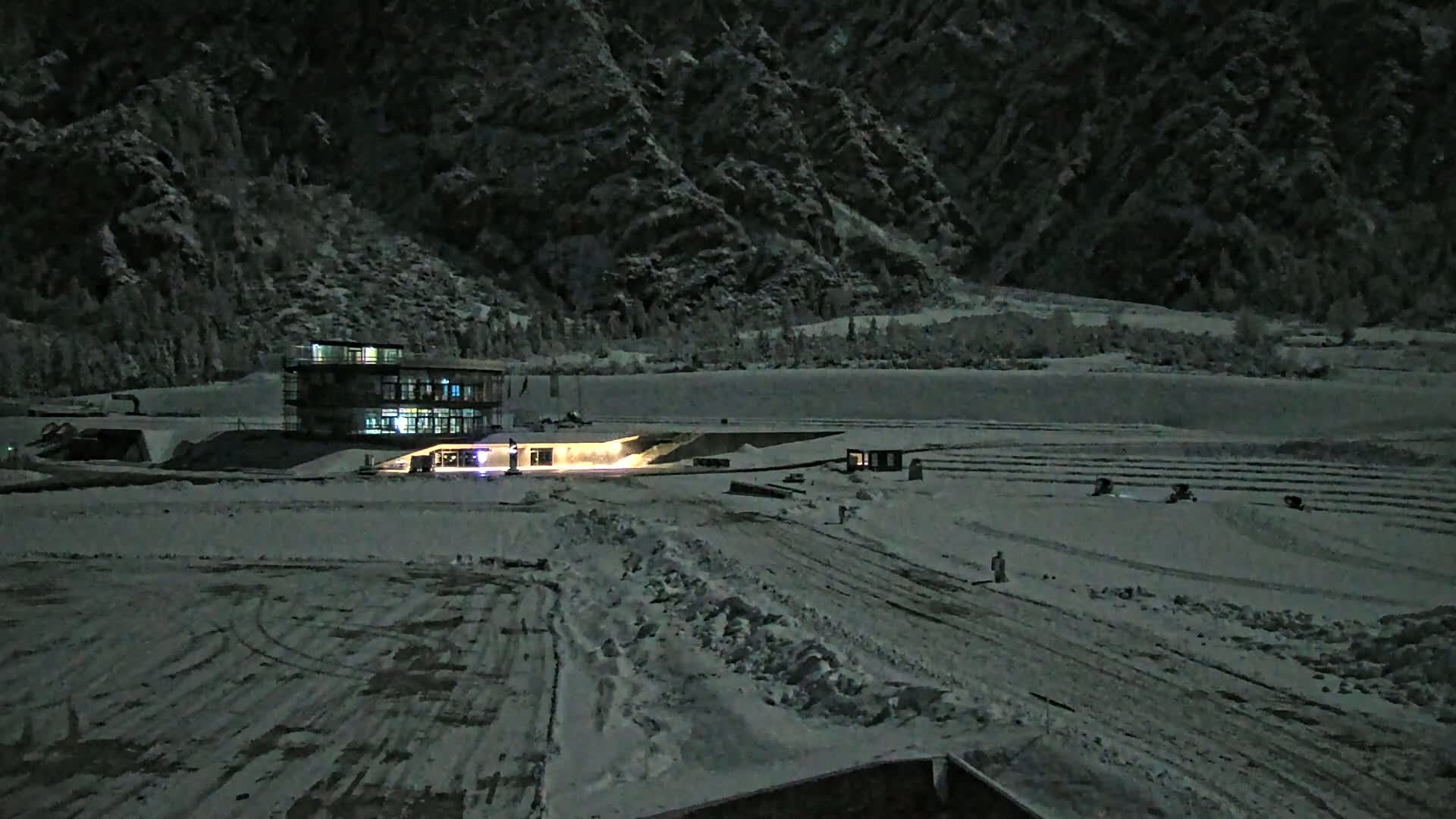 A snow-covered landscape at night, featuring a lit-up building near a flat, snow-covered area backed by snow-covered mountains.