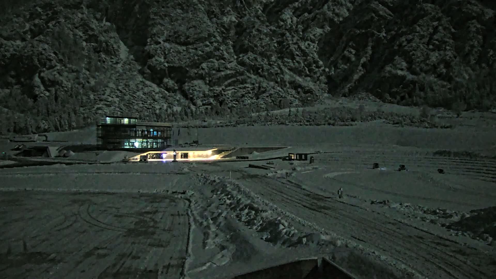 A snow-covered landscape at night, featuring a lit-up building near a mountain under a dark sky.