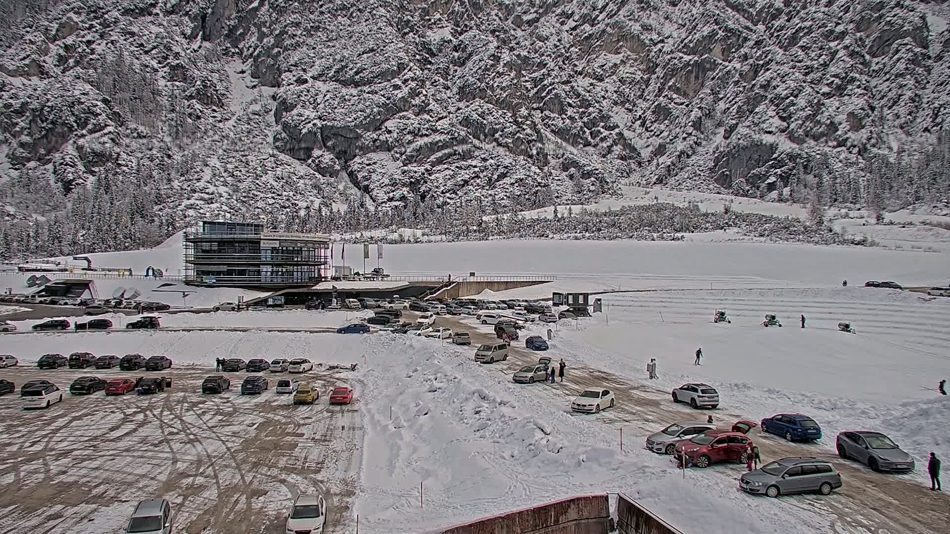 A snow-covered parking lot with numerous cars surrounds a modern building at the base of a snow-dusted mountain.