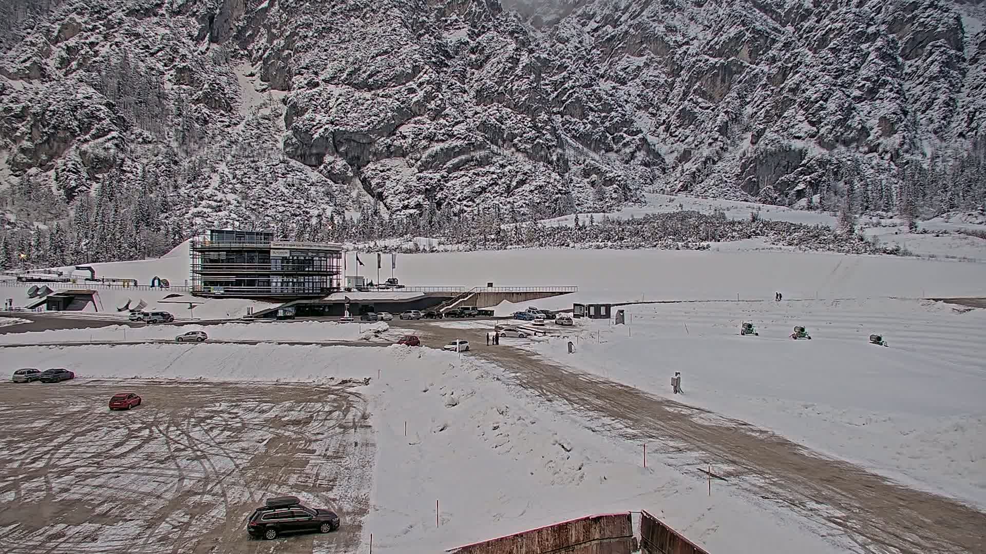 A snow-covered parking lot and roadway are situated in front of a modern building against a backdrop of snow-dusted, mountainous terrain.
