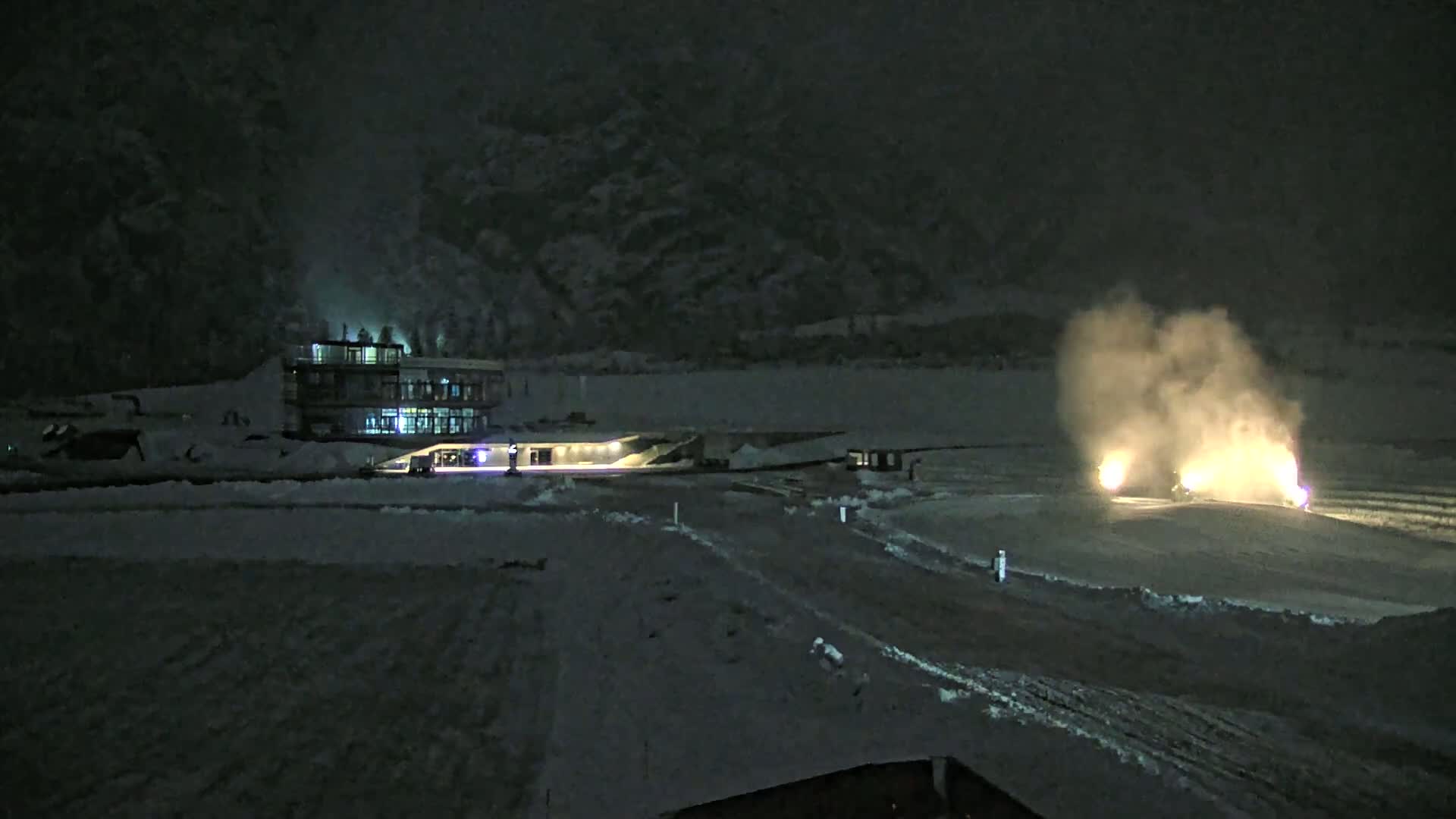 A snow-covered landscape at night, illuminated by a lit building and bright lights from vehicles emitting smoke in a field.