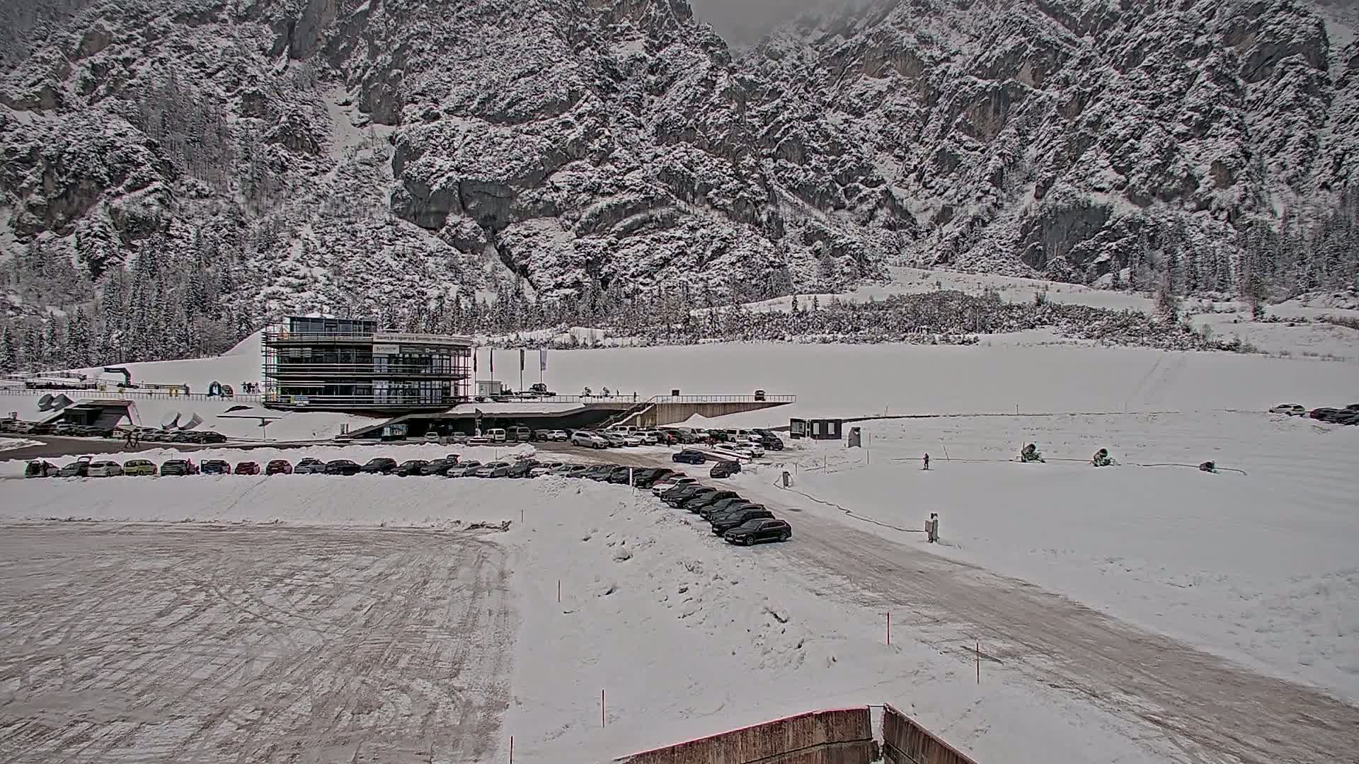 A snow-covered parking lot with cars and a modern building sits before a large, snow-dusted mountain range under an overcast sky.