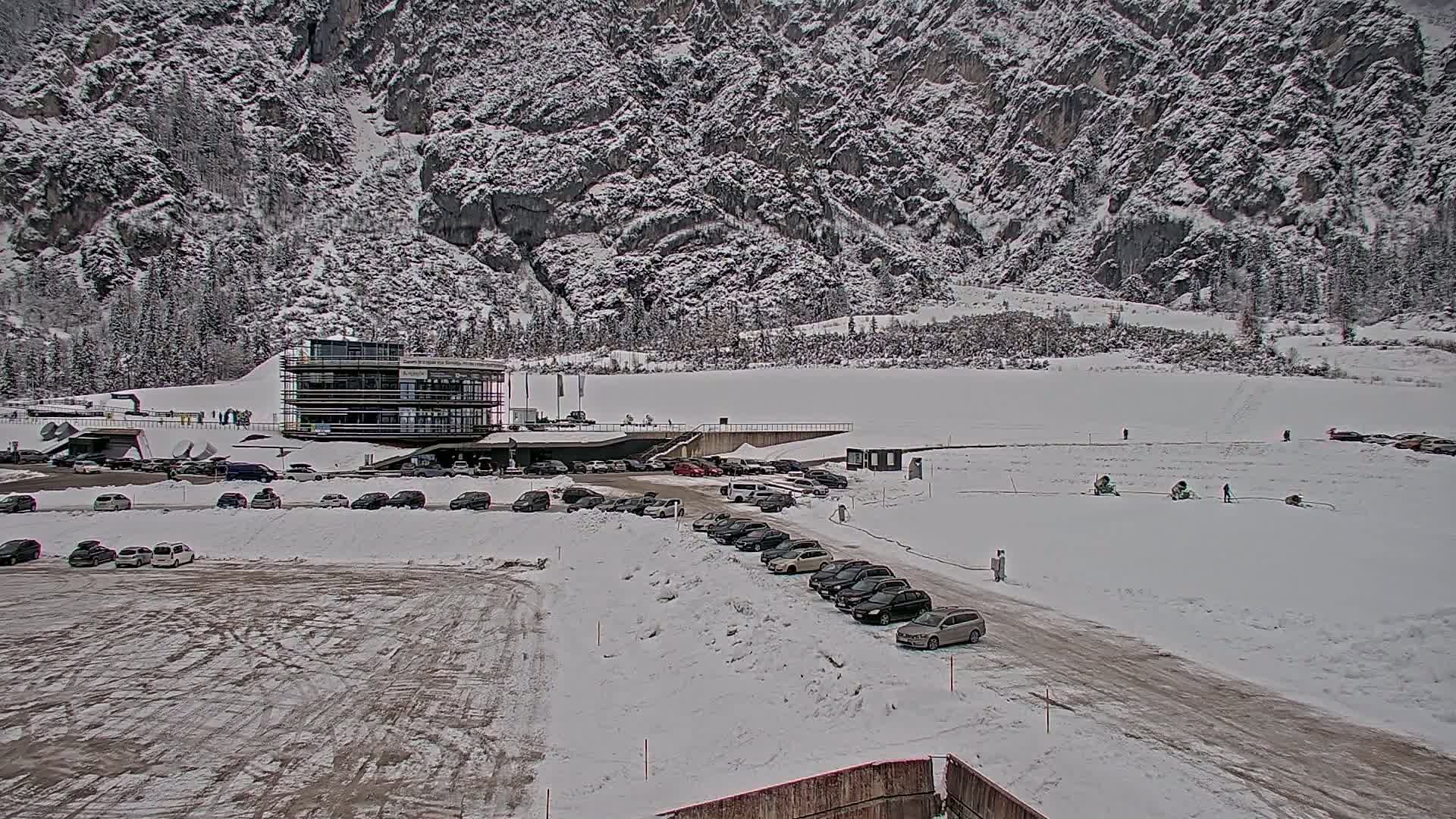 A snow-covered parking lot with cars, a multi-story building, and a large snow-covered field sits in front of a snow-dusted mountain.