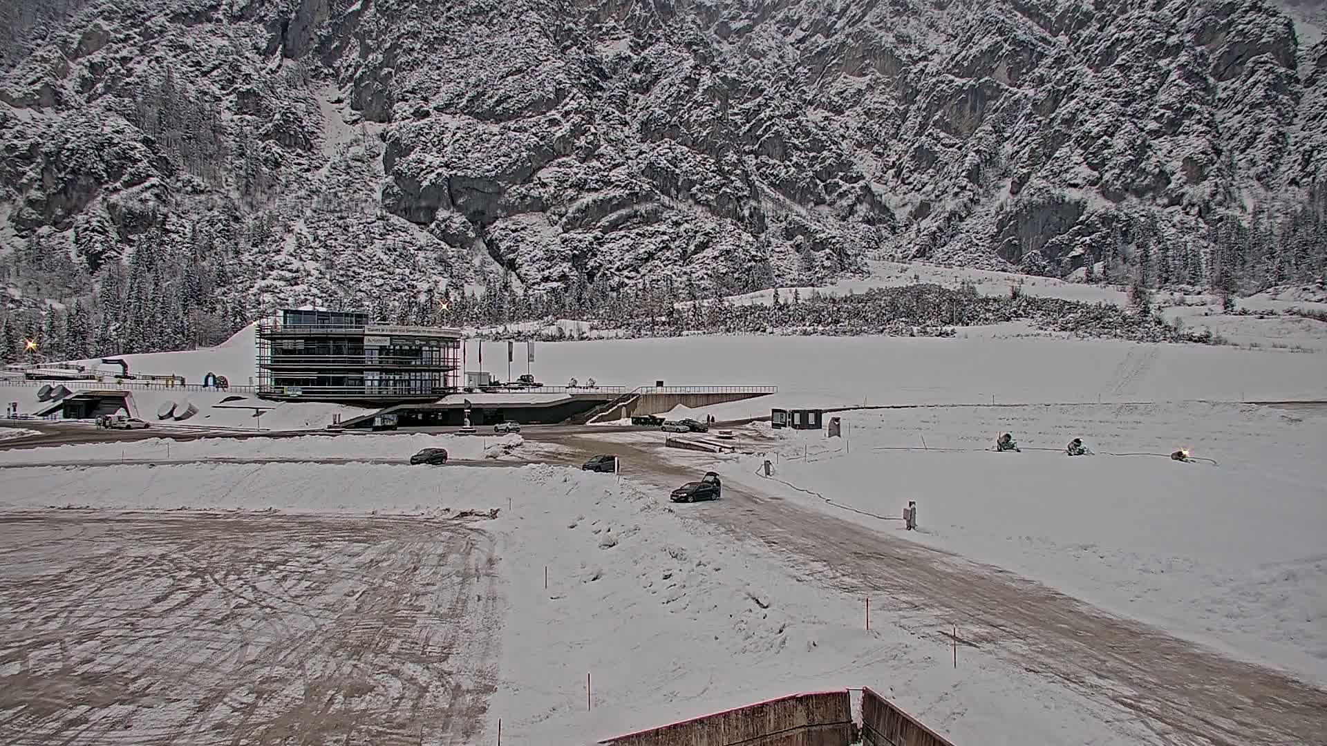 A snow-covered landscape with a modern building, several vehicles, and snowmobiles in the foreground, backed by a large, snow-dusted mountain.