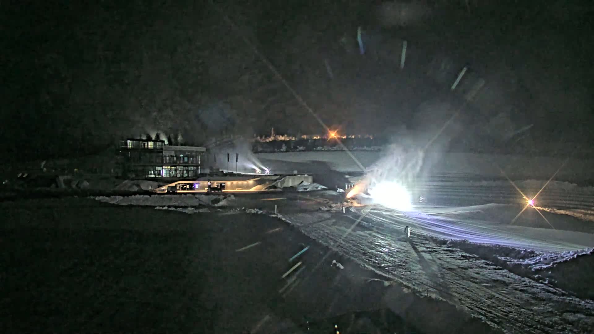 A snow-covered ski slope at night is illuminated by bright lights, with snowmaking machines in operation near a large building.