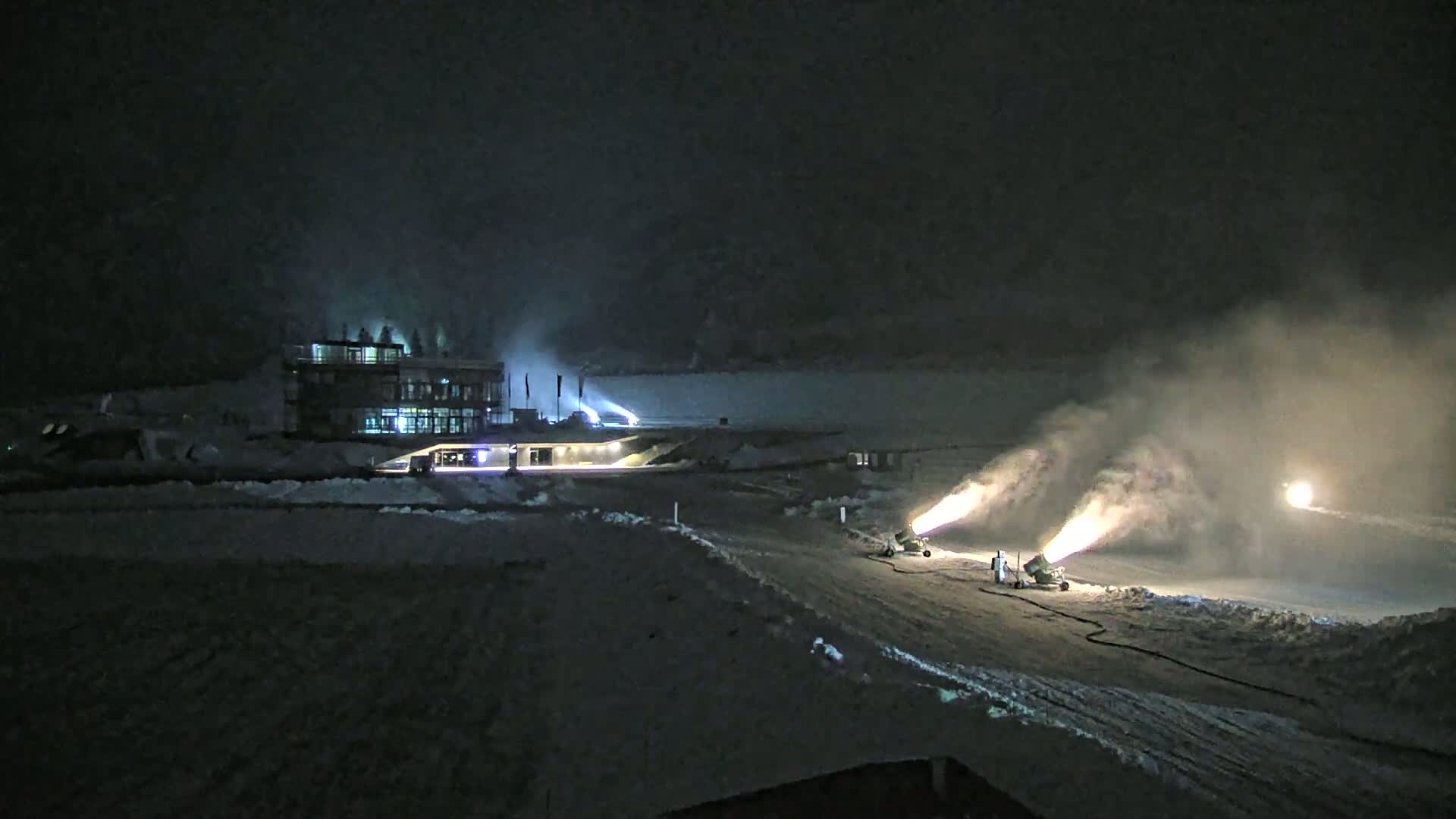 At night, two snow cannons emit plumes of white smoke onto a snow-covered landscape near a brightly lit building.