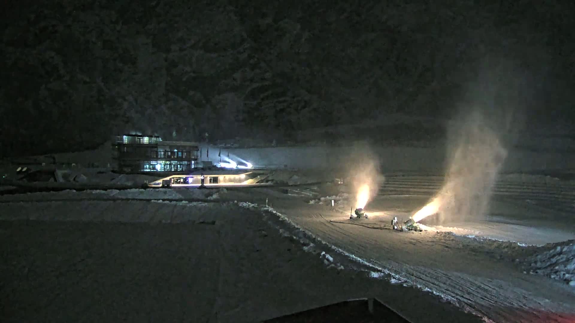 Two snow cannons are making snow on a snow-covered area at night, near a lit building and dark mountainside.