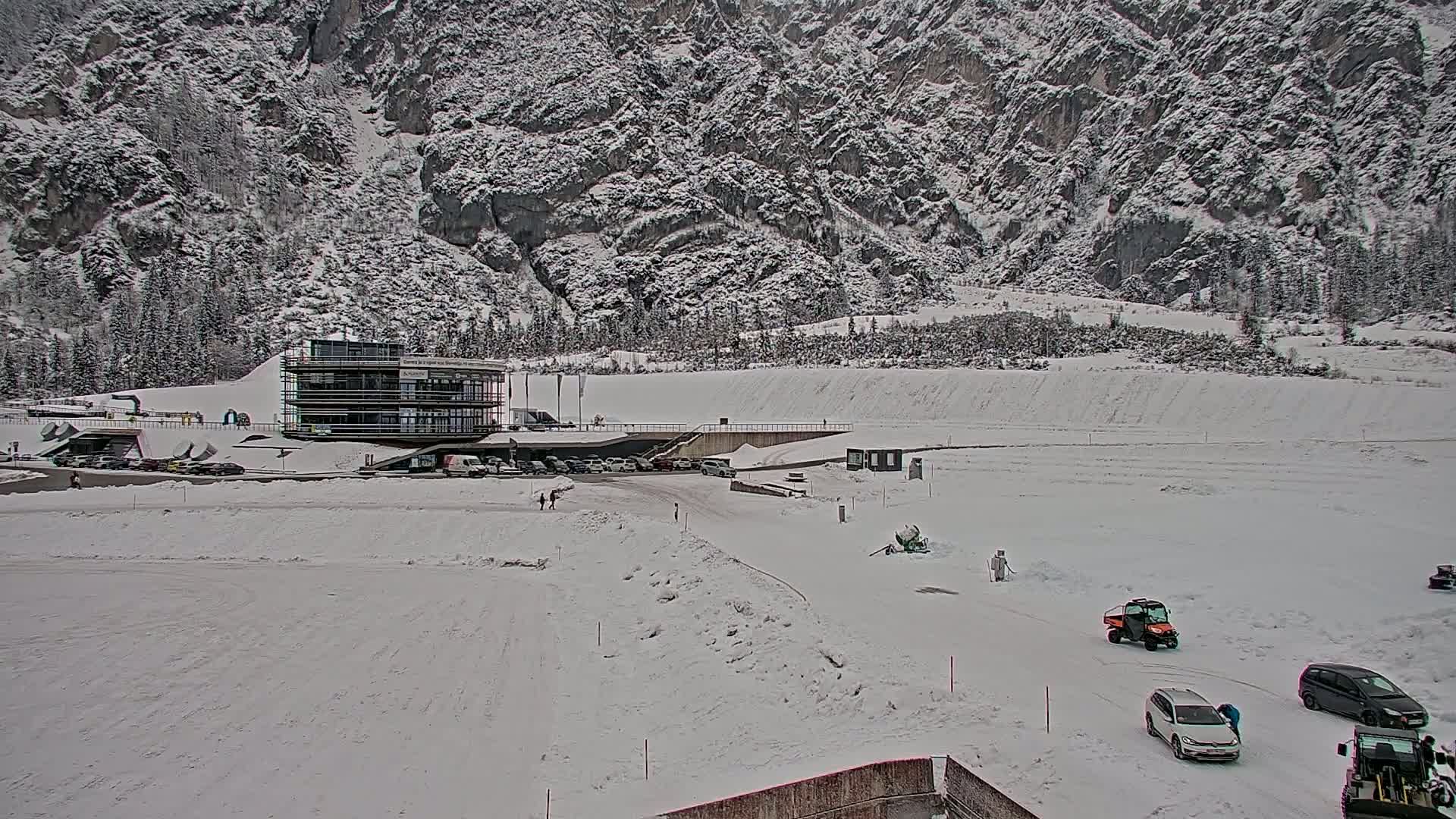 A snow-covered parking lot and modern building are situated at the base of a large, snow-dusted mountain.
