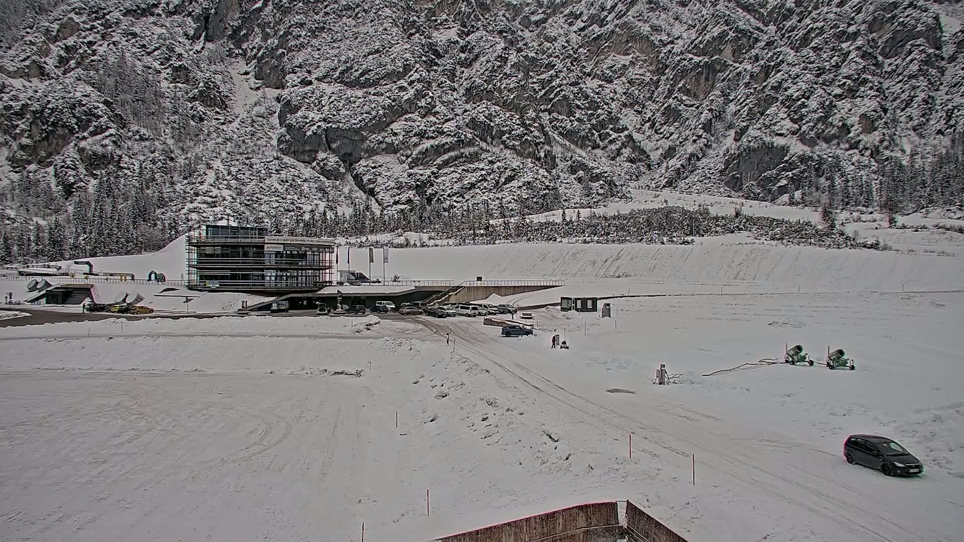 A snow-covered landscape features a modern building near a parking lot with several vehicles, and snow-making machines in the foreground, all set against a backdrop of a large, snow-dusted mountain.