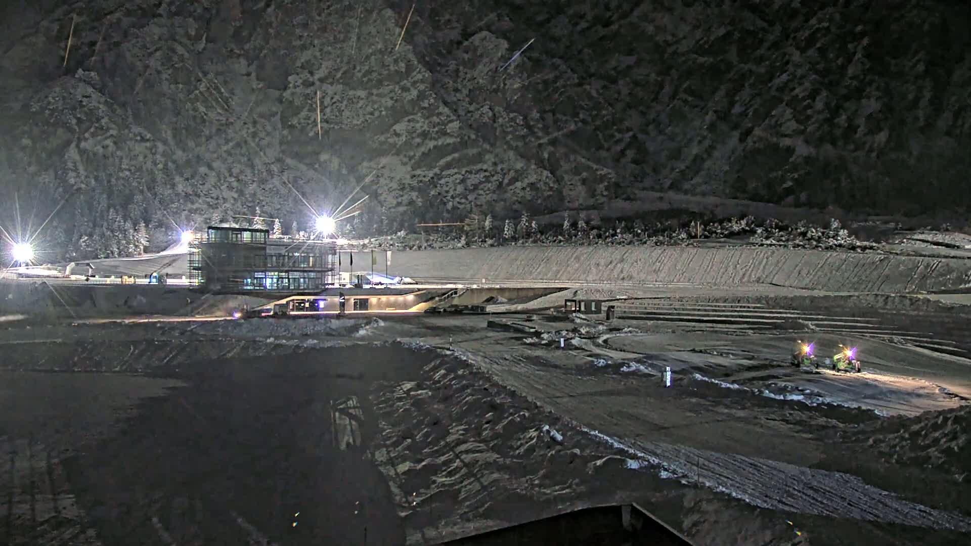 A snow-covered outdoor sports complex at night, illuminated by floodlights, with several small vehicles and a large building.
