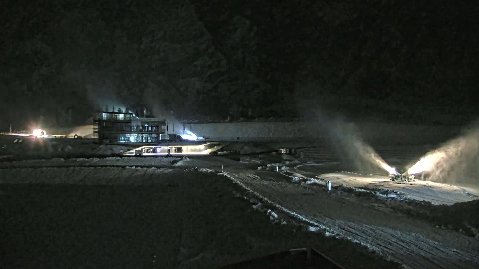 At night, two snow cannons are spraying snow onto a snow-covered area near a building, under dark, smoky conditions.