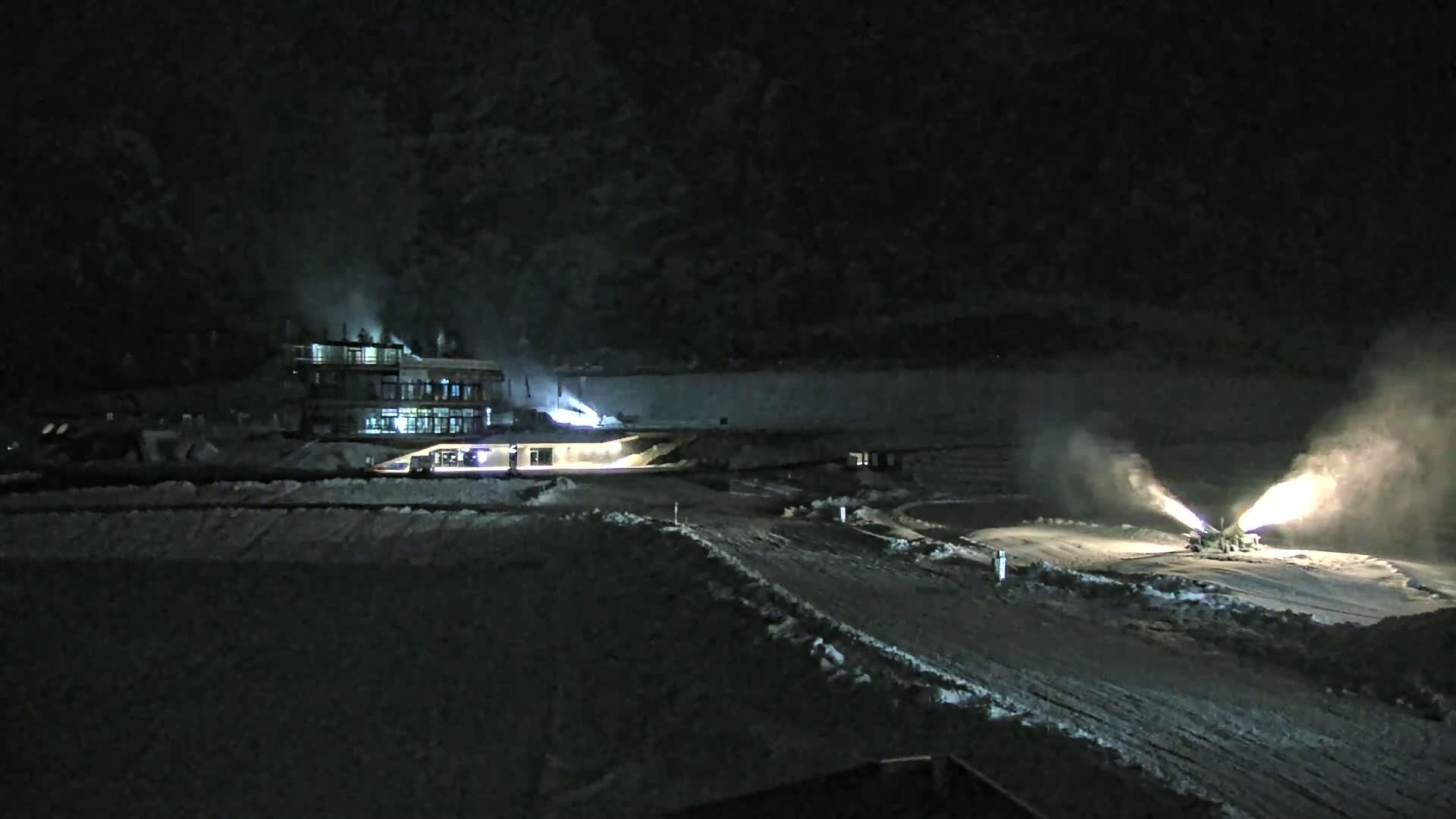 At night, snow cannons are making snow on a snowy landscape near a building.
