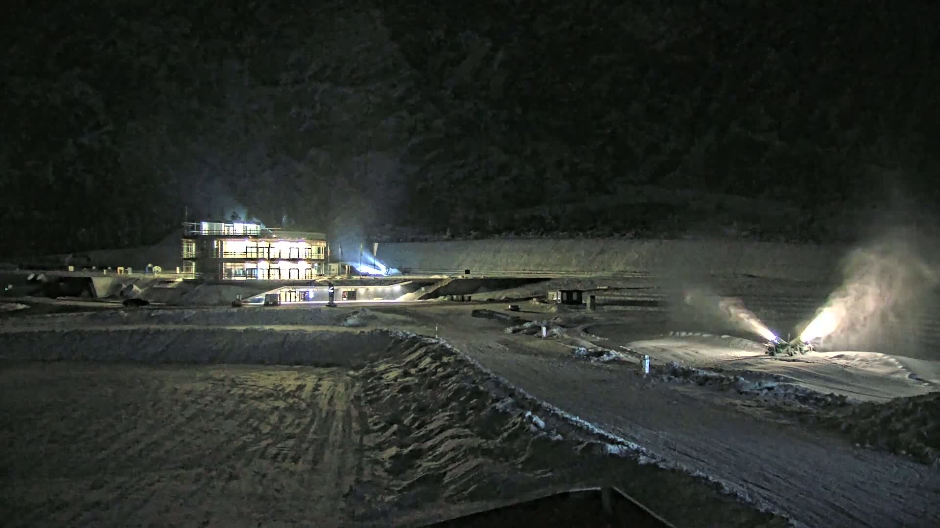 At night, snowmaking machines operate on a snow-covered area near a brightly lit building.