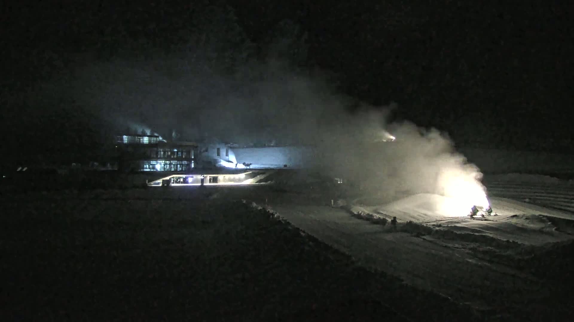 At night, snow cannons emit plumes of snow in a snowy area near a lit building.