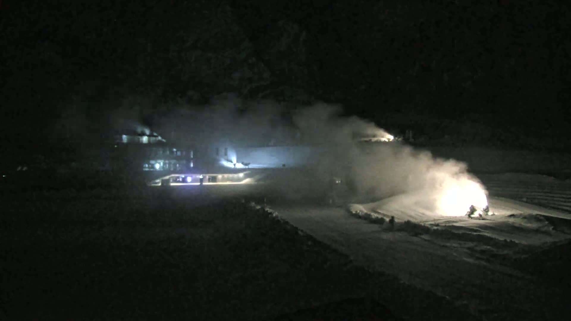 At night, snowmaking machines emit plumes of white vapor onto a snow-covered slope near a building.