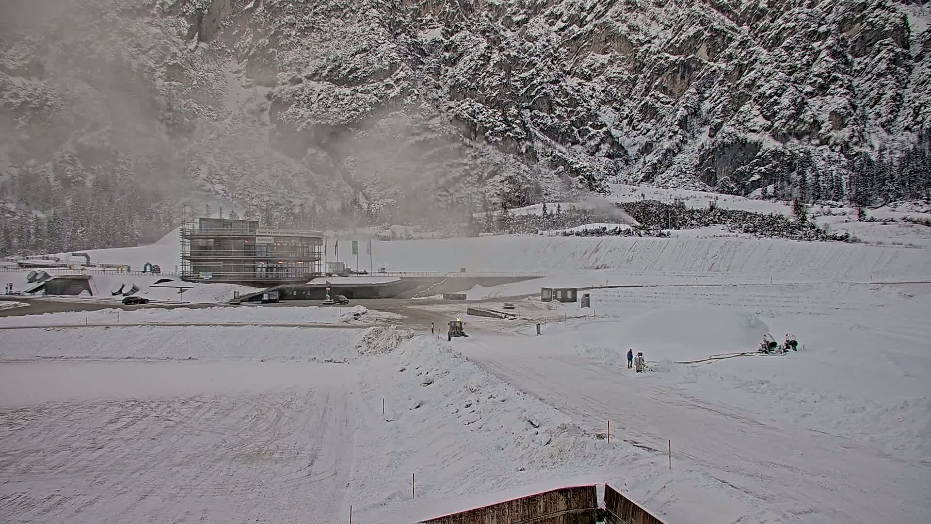A snow-covered landscape with a modern building, vehicles, and snow-making machines, set against a backdrop of a snow-laden mountain under misty conditions.