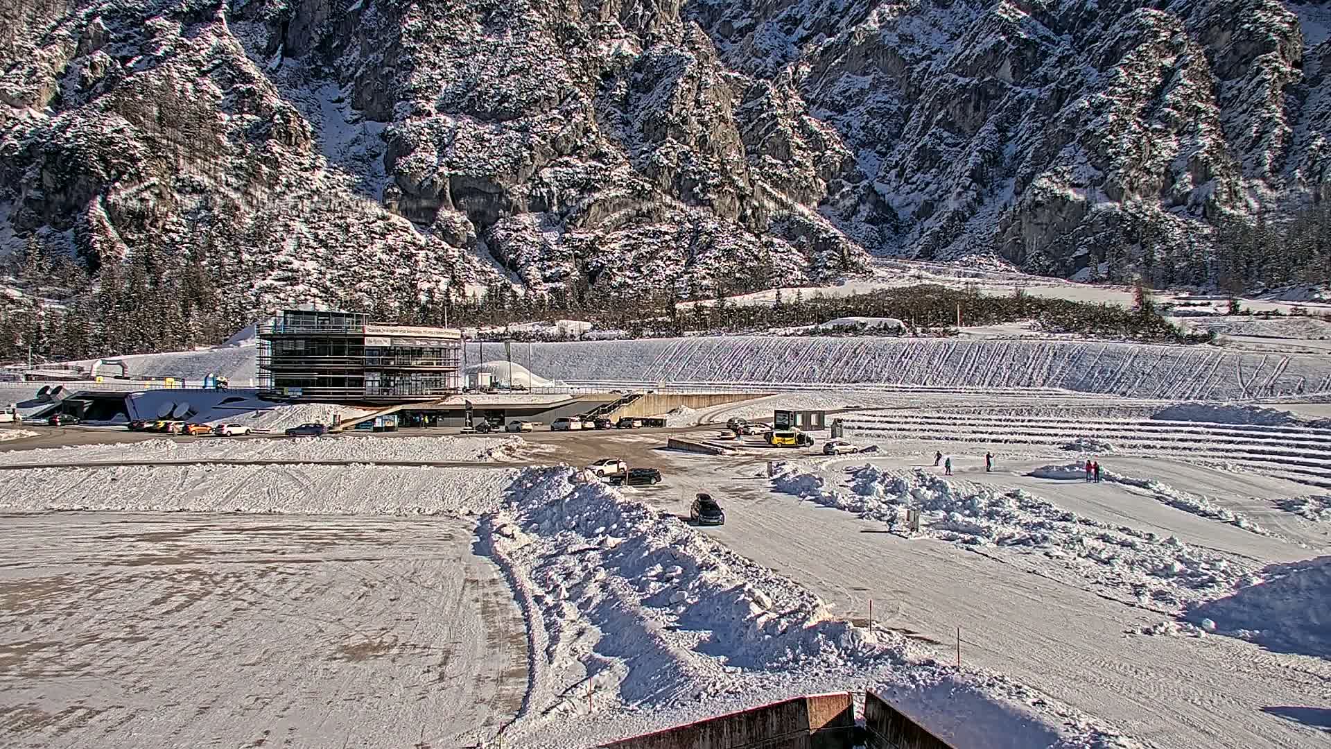 A snowy, winter scene shows a multi-level building near a parking lot and a large, snow-covered area with a few people, possibly enjoying winter activities, against the backdrop of a snow-dusted mountain range under a sunny sky.