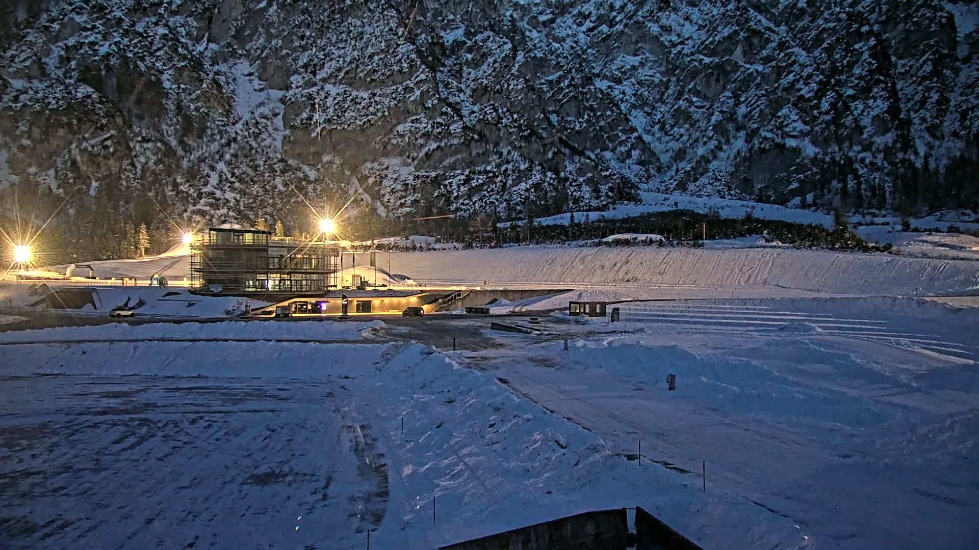 A snow-covered outdoor area with a lit building near a large, snow-dusted mountain at night.